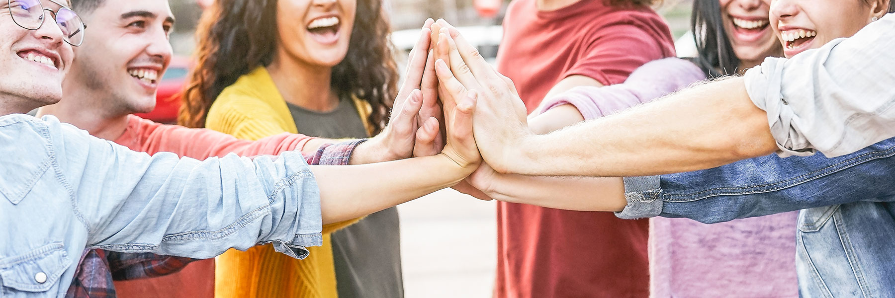 Group of seven smiling, diverse young adults with hands outstretched and touching in the center.