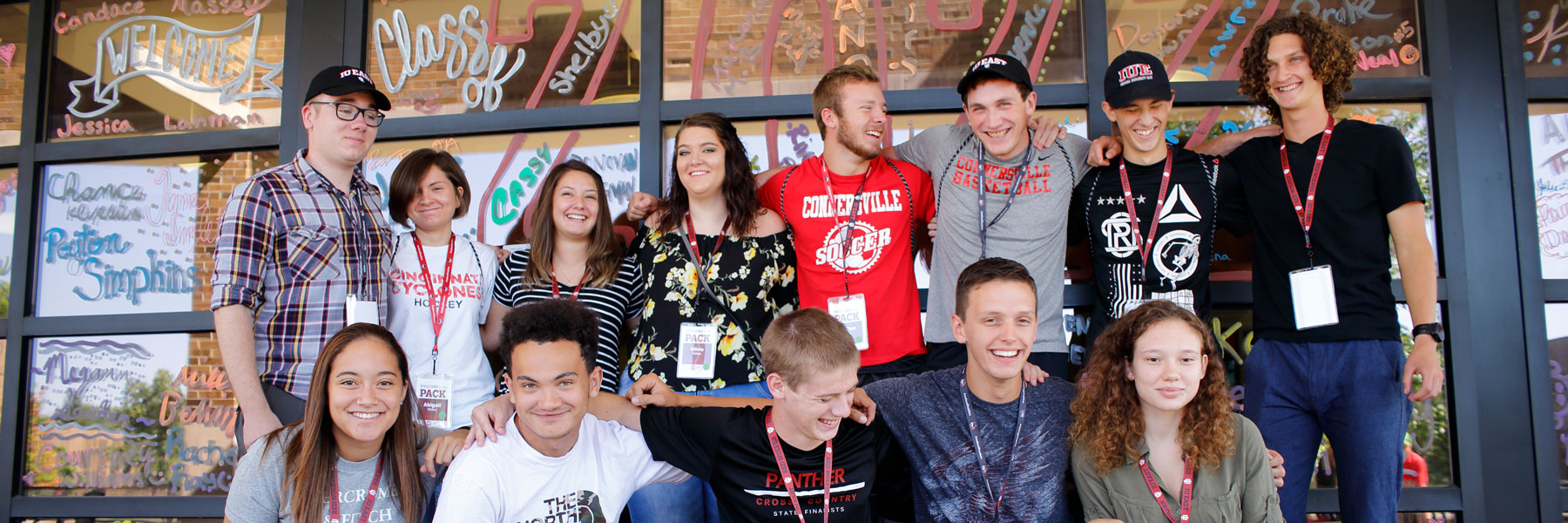 Group of diverse teens wearing id badges poses in front of a wall of windows decorated with art and graphics.