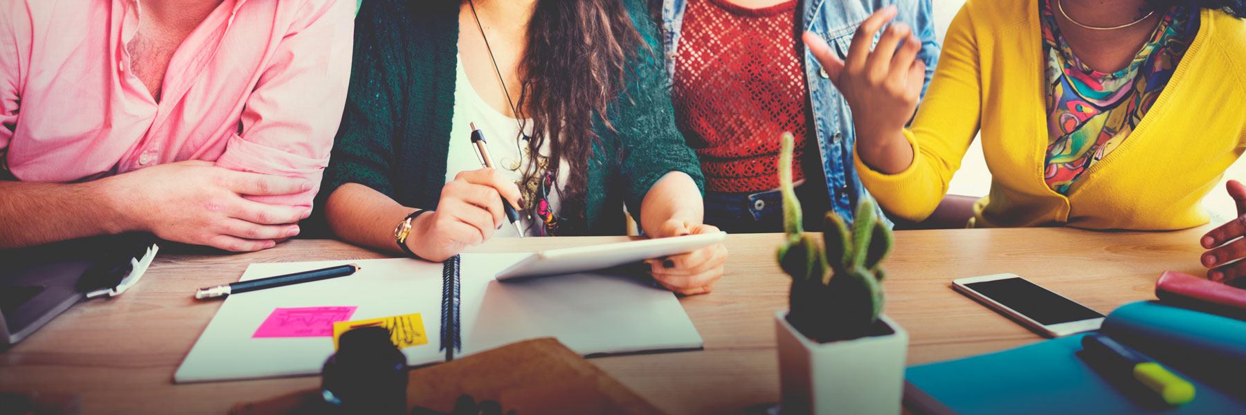 Torsos of six diverse young adults conversing and seated at a table with notebooks and mobile phones.