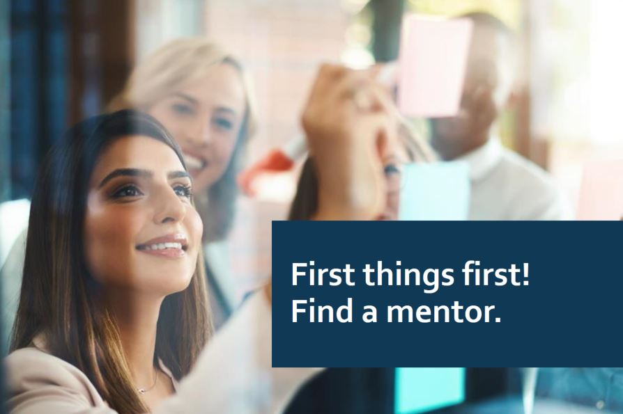 Young, smiling, professional woman with co-workers in background writes on sticky note affixed to window. Screen text says "First things first. Find a mentor!"