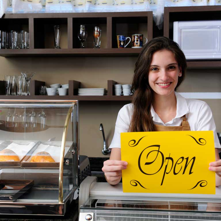 Young smiling woman behind a bakery counter. The sign she holds says Open.
